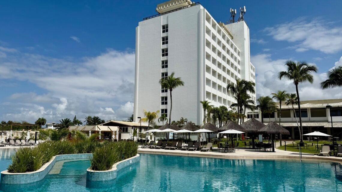 achada do hotel Deville Prime Salvador com coqueiros e área da piscina sob céu azul.