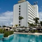 achada do hotel Deville Prime Salvador com coqueiros e área da piscina sob céu azul.