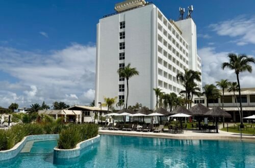 achada do hotel Deville Prime Salvador com coqueiros e área da piscina sob céu azul.