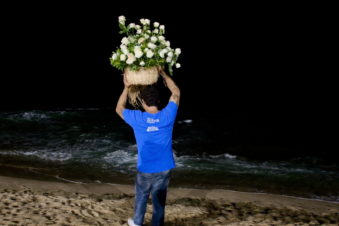 Homem de costas à beira-mar durante a noite, segurando um grande cesto de rosas brancas como oferenda para Iemanjá.