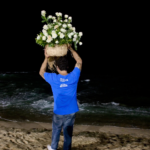 Homem de costas à beira-mar durante a noite, segurando um grande cesto de rosas brancas como oferenda para Iemanjá.