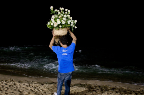 Homem de costas à beira-mar durante a noite, segurando um grande cesto de rosas brancas como oferenda para Iemanjá.