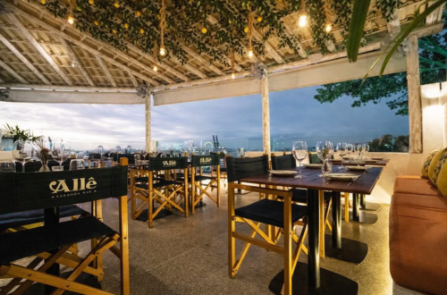 Vista interna da varanda do Allê Varanda Bar com mesas montadas e cadeiras de diretor pretas com a logomarca Allê. O teto rústico é decorado com limões sicilianos e iluminação quente, emoldurando a Baía de Todos-os-Santos ao fundo.