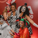 Margareth Menezes sorrindo com foliões fantasiados do bloco Os Mascarados.