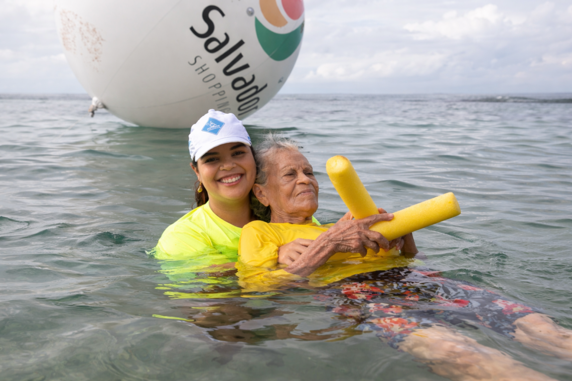 Voluntária do projeto ParaPraia sorri ao realizar banho de mar assistido em uma senhora idosa na Bacia das Moças, em Salvador. Ao fundo, balão promocional do Salvador Shopping.