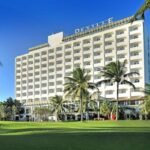 O Deville Prime Salvador exibe sua fachada com o nome do hotel visível, cercado por área verde e coqueiros.