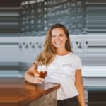 Débora Lehnen sorrindo com uma taça de cerveja na mão, representando a liderança da Proa Cervejaria.