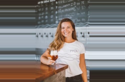 Débora Lehnen sorrindo com uma taça de cerveja na mão, representando a liderança da Proa Cervejaria.