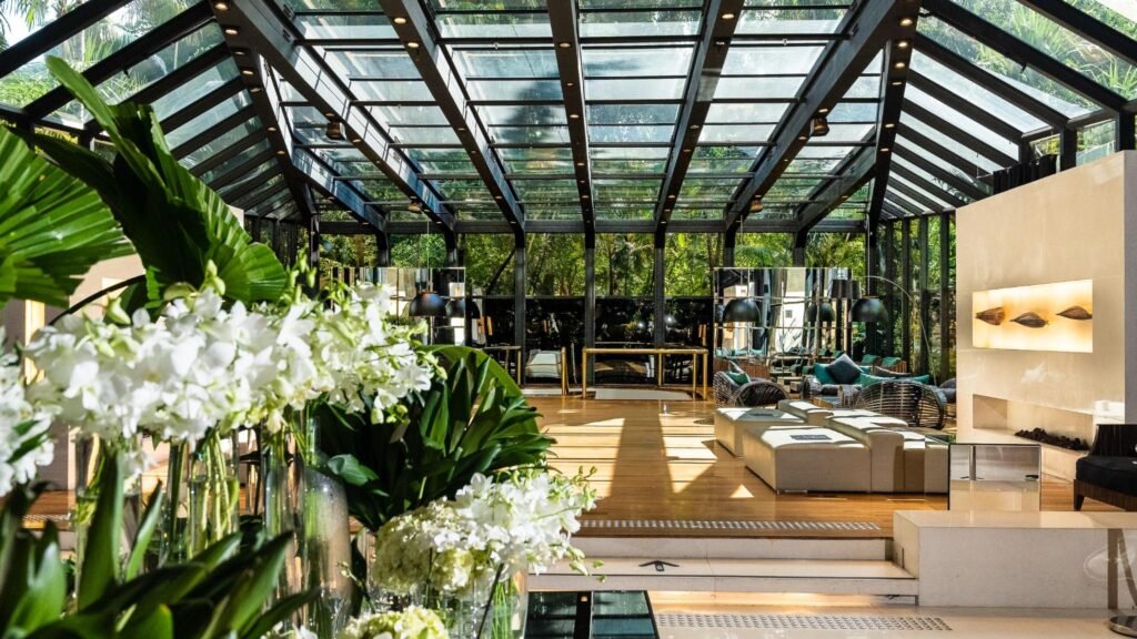 Vista do saguão do Tivoli Mofarrej São Paulo com sua icônica estrutura de vidro e aço. O espaço é iluminado por luz natural que atravessa a claraboia e revela a vegetação externa. No primeiro plano, arranjos de flores brancas e folhagens tropicais. O piso de madeira e os móveis de design completam o ambiente.