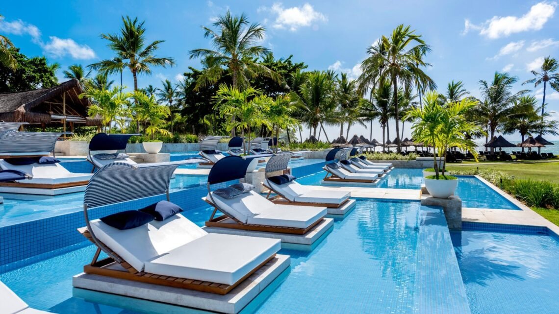 oto da piscina do Tivoli Ecoresort Praia do Forte, na Bahia. Em primeiro plano, espreguiçadeiras com colchões brancos, travesseiros azuis e coberturas listradas de azul e branco estão posicionadas dentro da água da piscina. Ao fundo, uma área com gramado, coqueiros e quiosques de palha dá para o mar sob um céu azul.