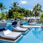 oto da piscina do Tivoli Ecoresort Praia do Forte, na Bahia. Em primeiro plano, espreguiçadeiras com colchões brancos, travesseiros azuis e coberturas listradas de azul e branco estão posicionadas dentro da água da piscina. Ao fundo, uma área com gramado, coqueiros e quiosques de palha dá para o mar sob um céu azul.