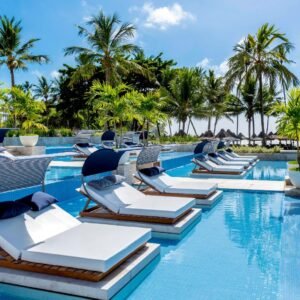 oto da piscina do Tivoli Ecoresort Praia do Forte, na Bahia. Em primeiro plano, espreguiçadeiras com colchões brancos, travesseiros azuis e coberturas listradas de azul e branco estão posicionadas dentro da água da piscina. Ao fundo, uma área com gramado, coqueiros e quiosques de palha dá para o mar sob um céu azul.