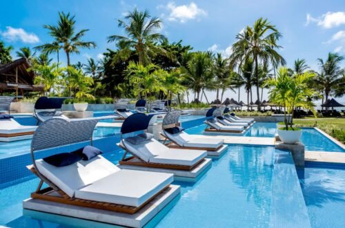 oto da piscina do Tivoli Ecoresort Praia do Forte, na Bahia. Em primeiro plano, espreguiçadeiras com colchões brancos, travesseiros azuis e coberturas listradas de azul e branco estão posicionadas dentro da água da piscina. Ao fundo, uma área com gramado, coqueiros e quiosques de palha dá para o mar sob um céu azul.