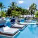 oto da piscina do Tivoli Ecoresort Praia do Forte, na Bahia. Em primeiro plano, espreguiçadeiras com colchões brancos, travesseiros azuis e coberturas listradas de azul e branco estão posicionadas dentro da água da piscina. Ao fundo, uma área com gramado, coqueiros e quiosques de palha dá para o mar sob um céu azul.