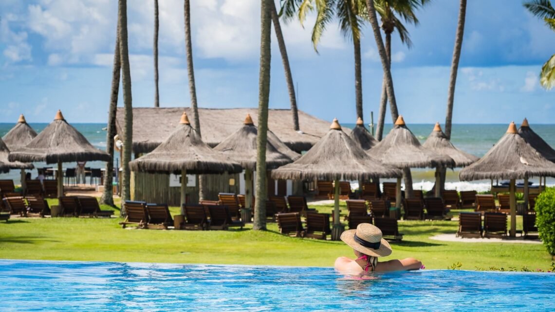 Mulher com chapéu de palha relaxando em piscina de borda infinita de resort na Praia do Forte com coqueiros e vista para o mar.