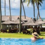 Mulher com chapéu de palha relaxando em piscina de borda infinita de resort na Praia do Forte com coqueiros e vista para o mar.