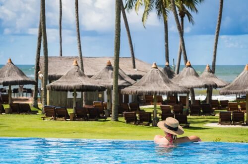 Mulher com chapéu de palha relaxando em piscina de borda infinita de resort na Praia do Forte com coqueiros e vista para o mar.