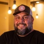 Sandro Borges, chef convidado do PorcoDay Reconvexo, com boné e camiseta preta em ambiente de restaurante.