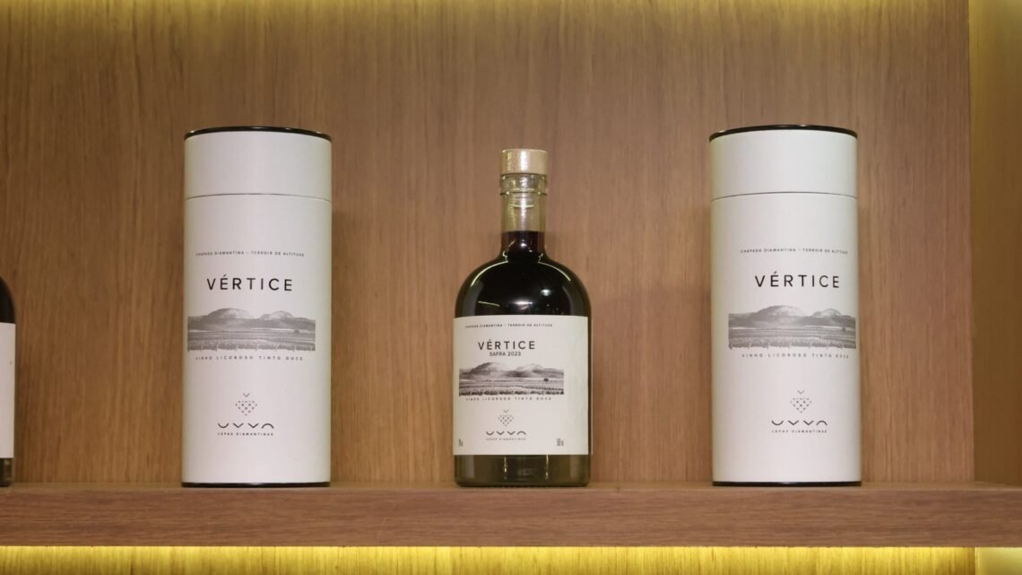 Garrafa do Vértice, primeiro vinho licoroso da UVVA, entre duas embalagens em uma prateleira de madeira.