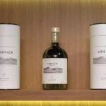 Garrafa do Vértice, primeiro vinho licoroso da UVVA, entre duas embalagens em uma prateleira de madeira.