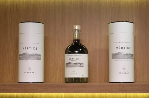 Garrafa do Vértice, primeiro vinho licoroso da UVVA, entre duas embalagens em uma prateleira de madeira.
